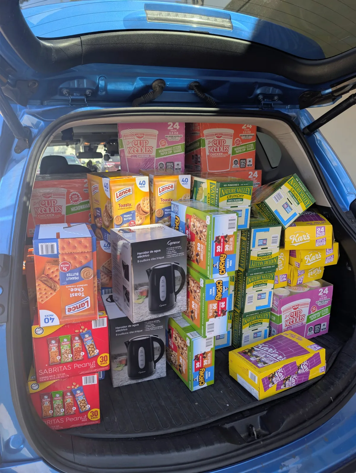 A car boot packed with cases of snacks — Cup Noodles, Lance crackers, Nature Valley and Chewy granola bars, trail mix and electric kettles.